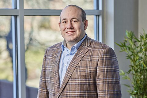 A man in a brown plaid blazer and light blue shirt stands indoors near a window, smiling at the camera with greenery visible outside.