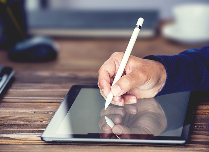 A hand using a stylus to write or draw on a tablet placed on a wooden desk.