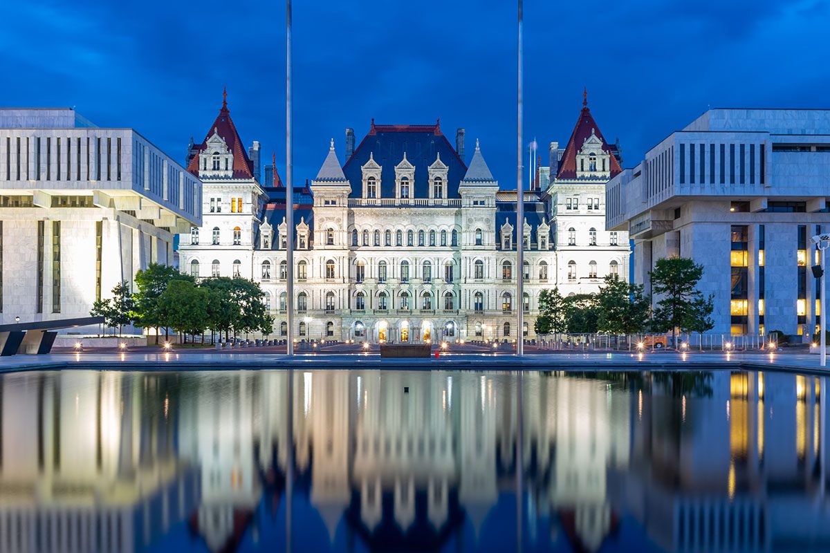 The New York State Capitol building at dusk, illuminated and reflected in a large, still pool, flanked by modern office buildings.