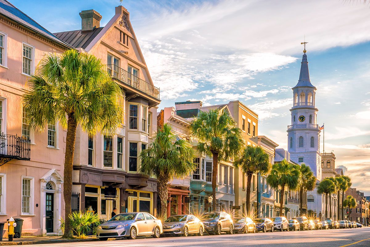 A row of historic buildings with palm trees and parked cars lines a sunny street; a church steeple rises in the background.