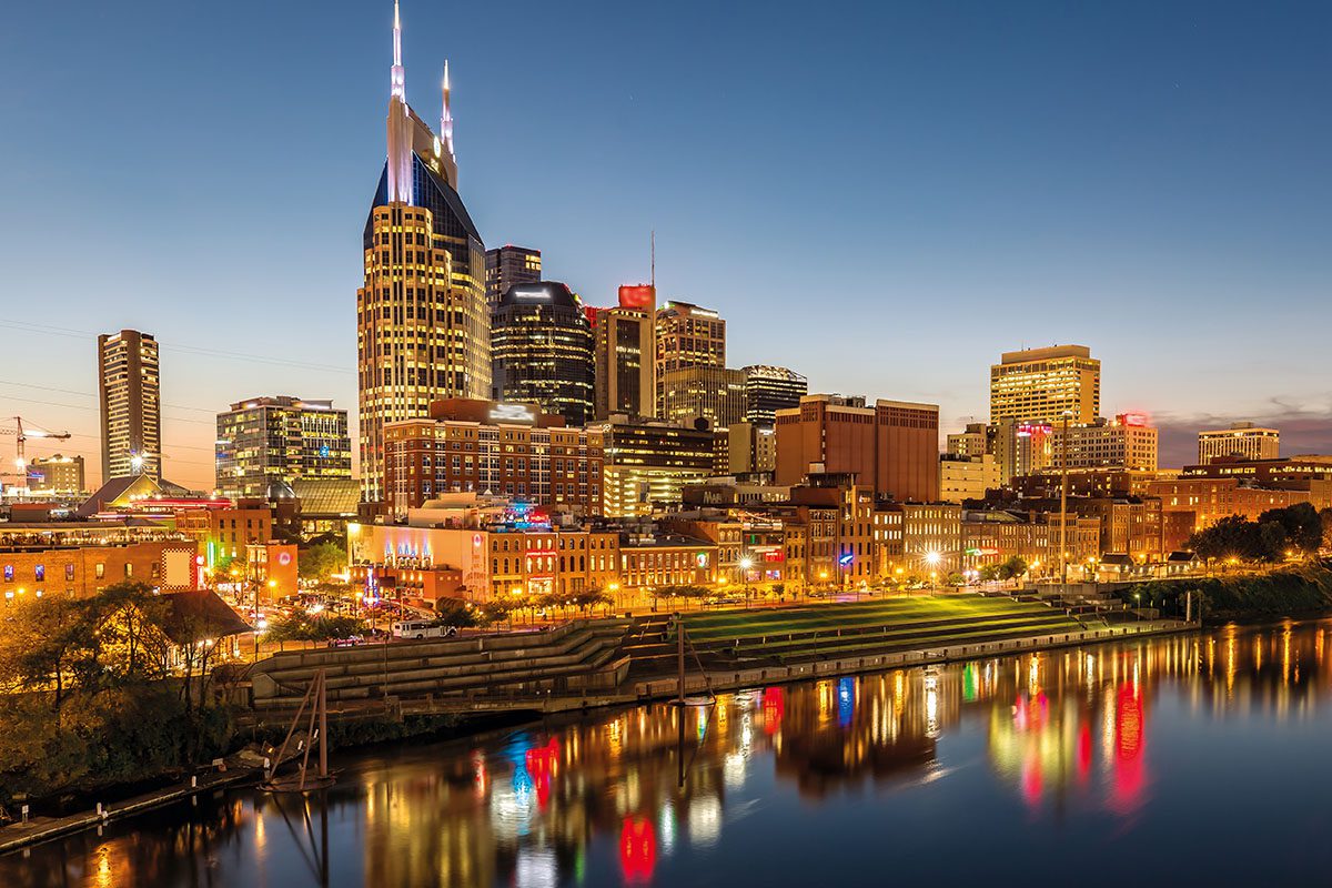 Nashville city skyline at dusk with illuminated buildings reflecting on the river in the foreground.