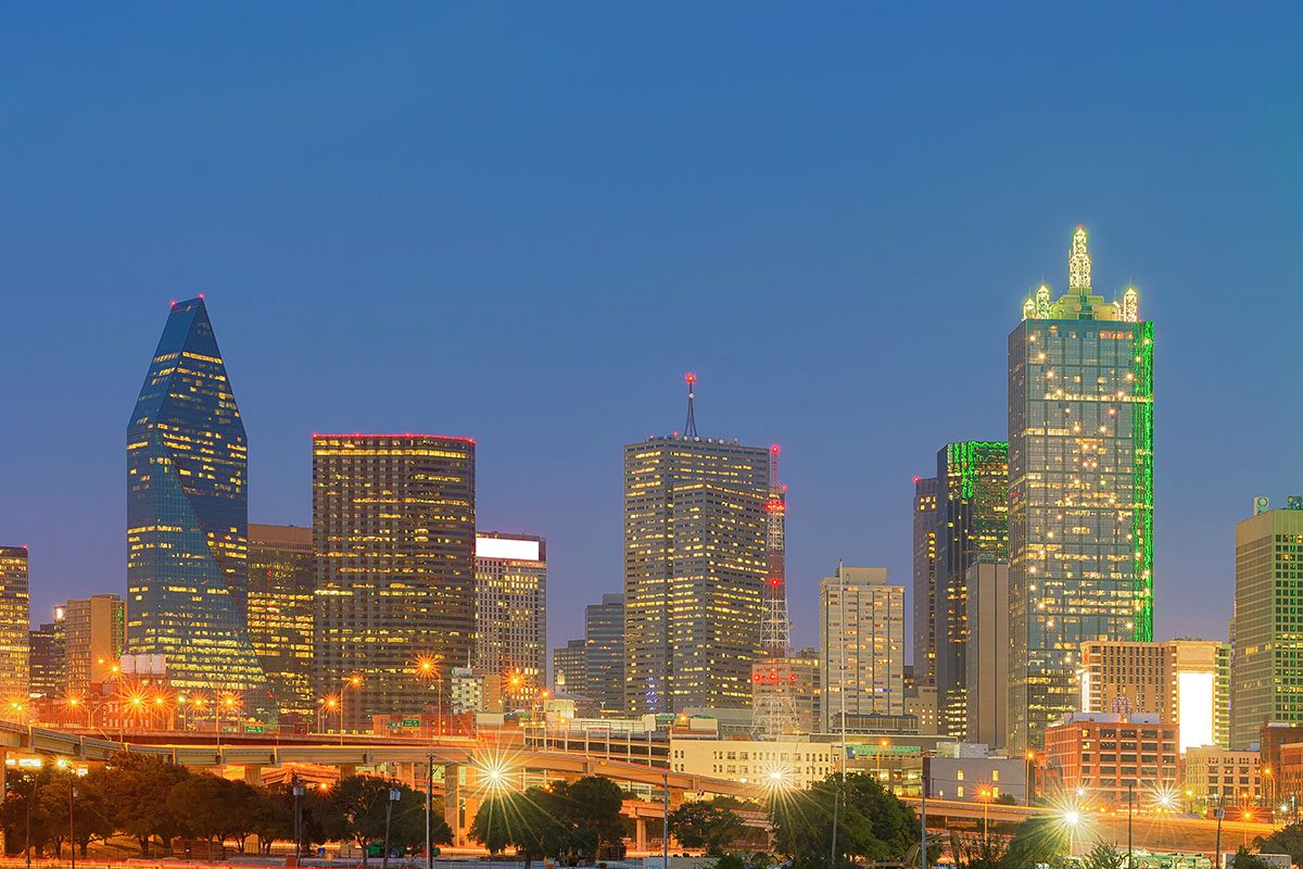 Dallas city skyline at dusk with illuminated skyscrapers and an overpass in the foreground.