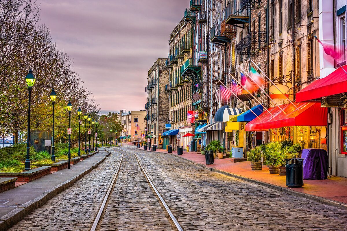 A cobblestone street lined with colorful awnings, flags, street lamps, and historic buildings next to leafless trees under a cloudy sky.