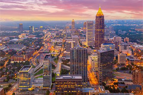 Aerial view of a cityscape at sunset with tall skyscrapers, illuminated streets, and a colorful sky.