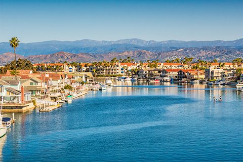 A waterfront residential neighborhood with houses along a calm canal, boats docked by the shore, and mountains visible in the background under a clear sky.