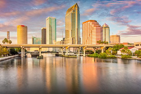 Downtown city skyline with modern high-rise buildings reflected in a calm river at sunset, with a bridge crossing the water in the foreground.