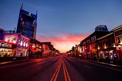 Empty street in downtown Nashville at sunset, with neon signs and buildings lit on both sides, and a tall skyscraper in the background.