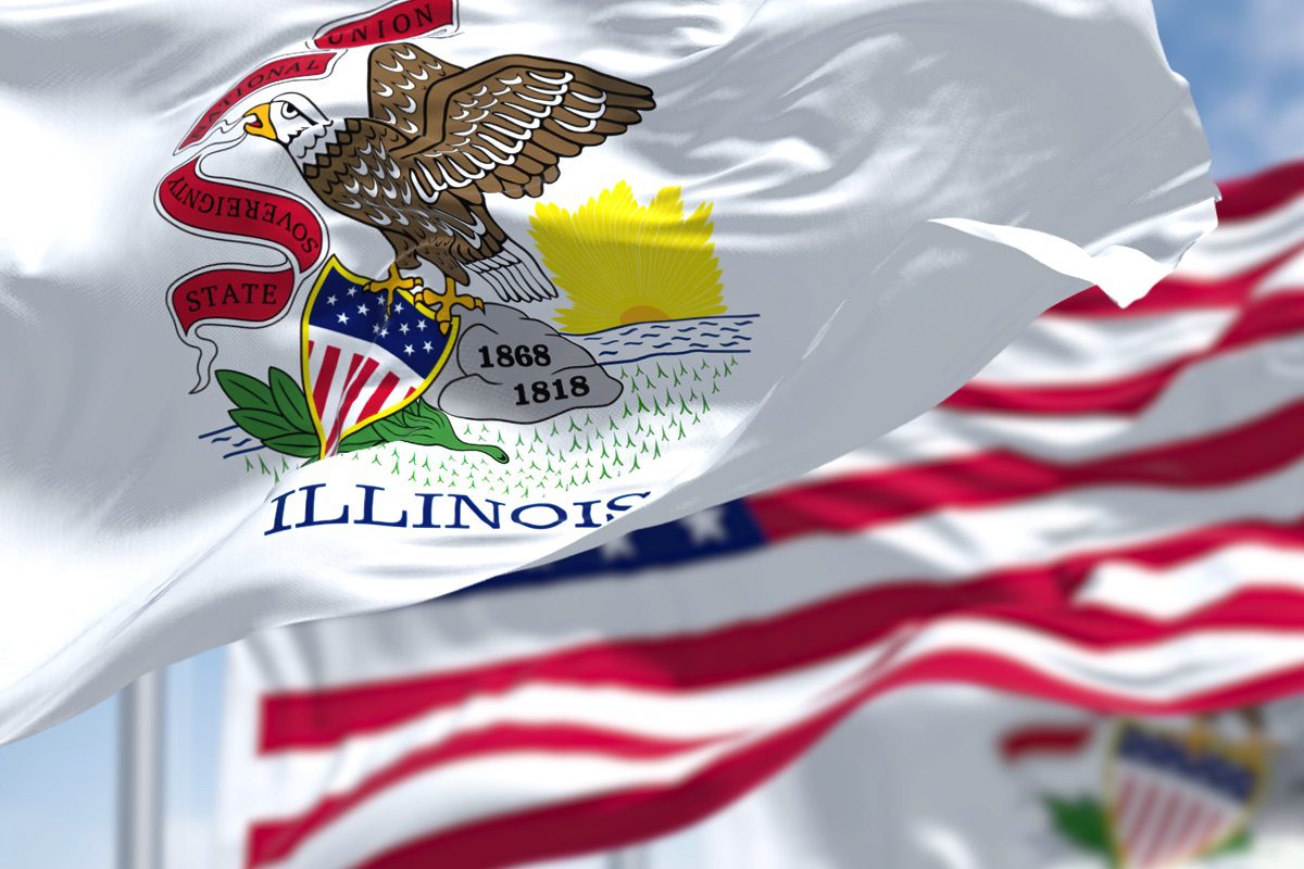 The Illinois state flag, featuring an eagle holding a shield and a banner with the state motto, waves in the foreground with another flag visible in the background.