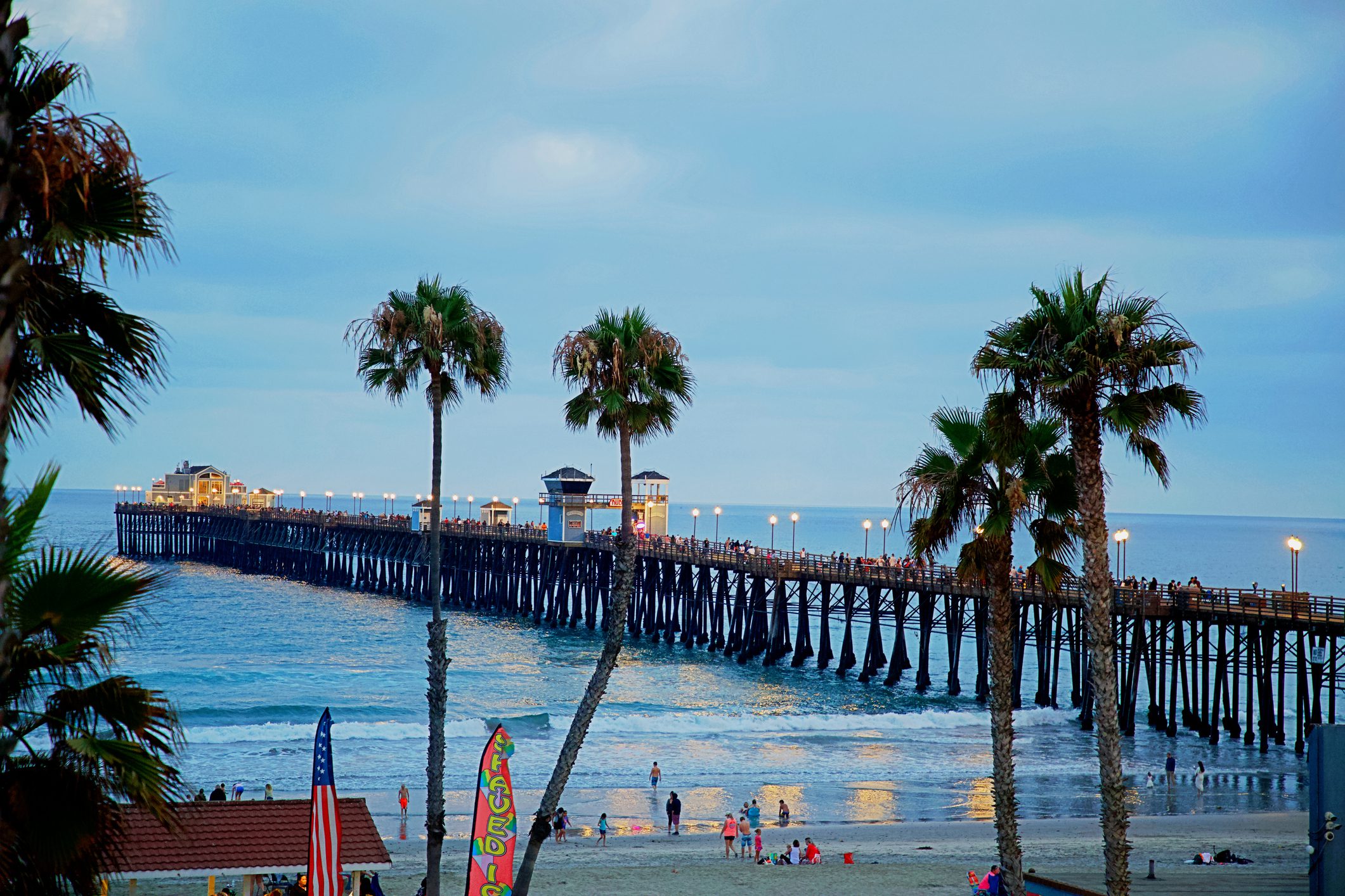 A long wooden pier stretches into the ocean at dusk, lined with lit lamps, palm trees in the foreground, and people on the beach and pier.