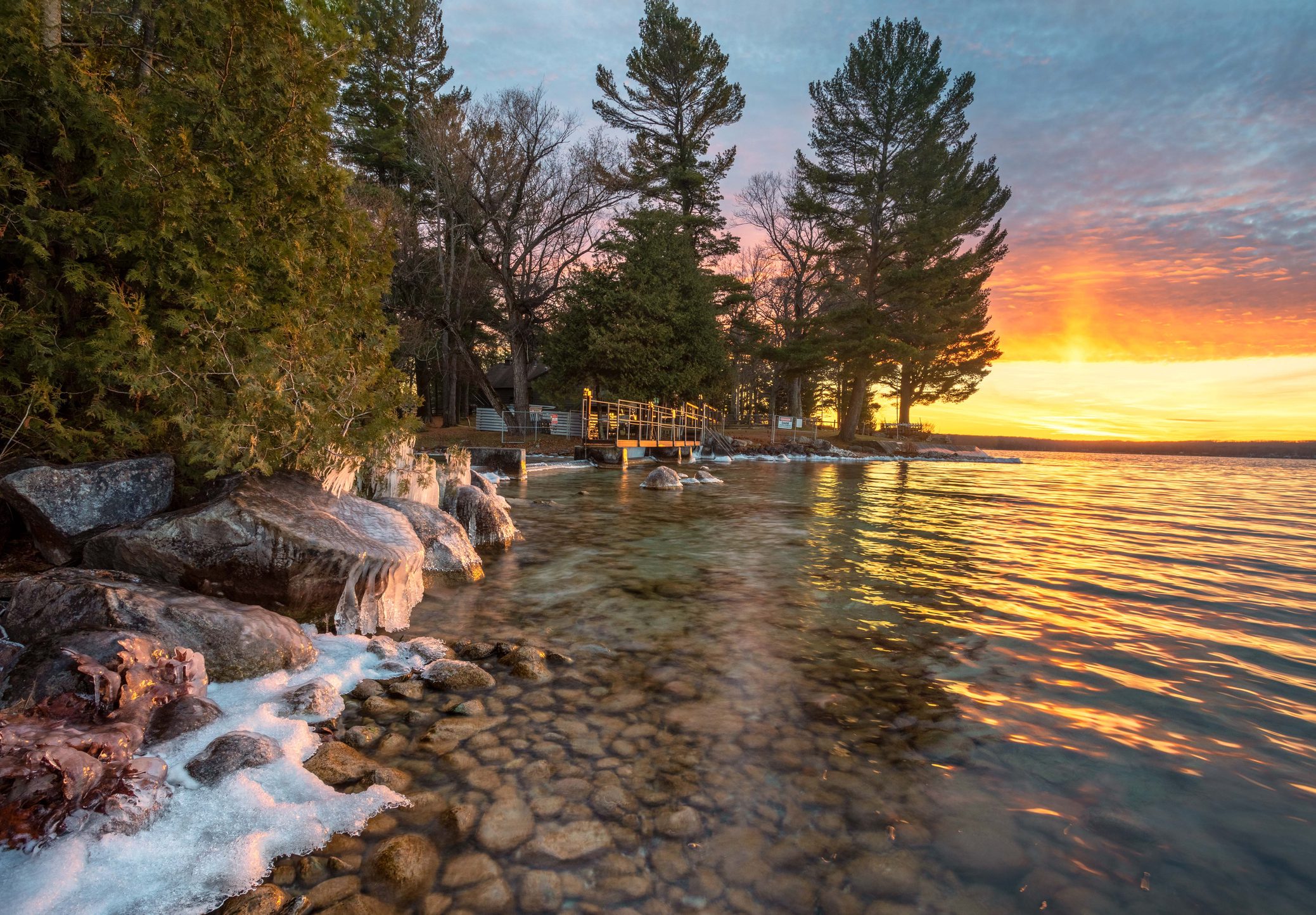 Rocks along a lakeshore with some ice, clear water, and a pier; trees line the shore and the sun sets in the background, casting an orange glow.