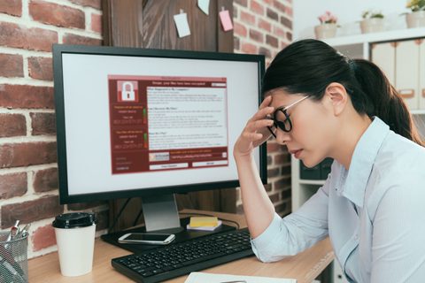 A woman sits at a desk, looking stressed, with her hand on her forehead. A computer screen in front of her displays a ransomware warning message.