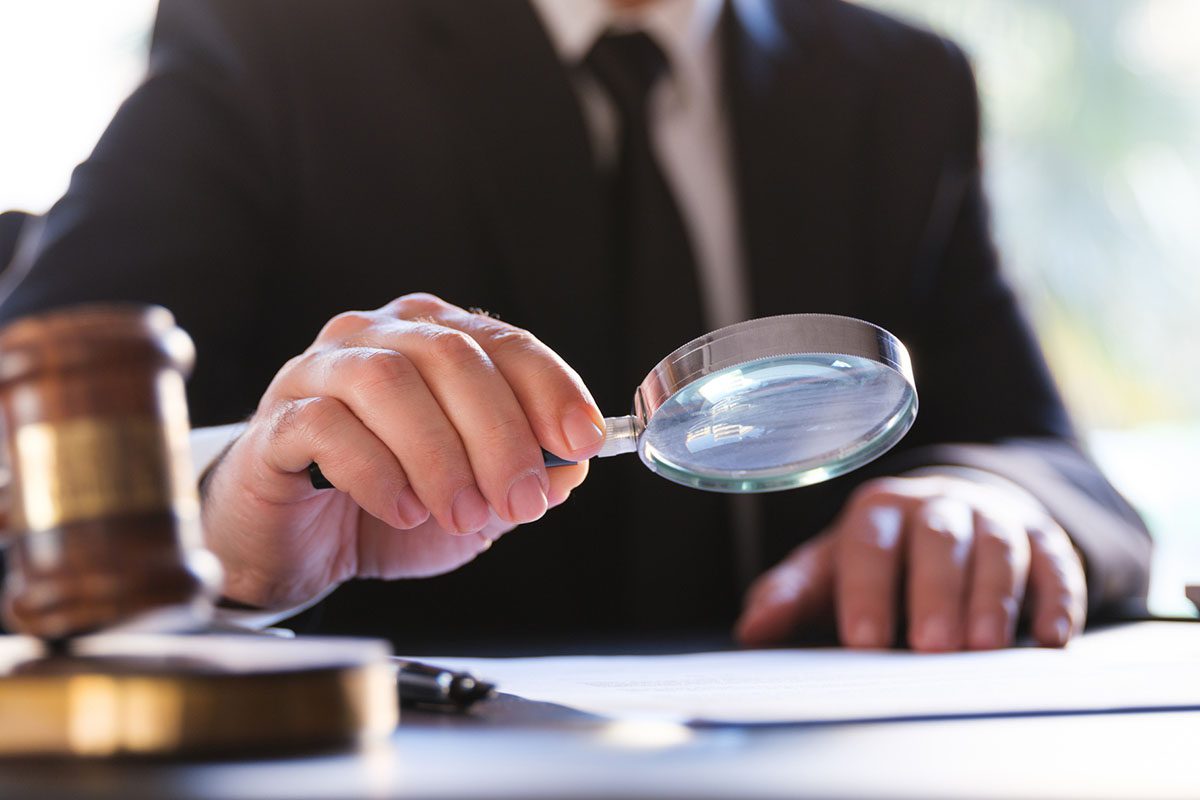 A person in a suit examines a document with a magnifying glass while sitting at a desk with a gavel nearby.
