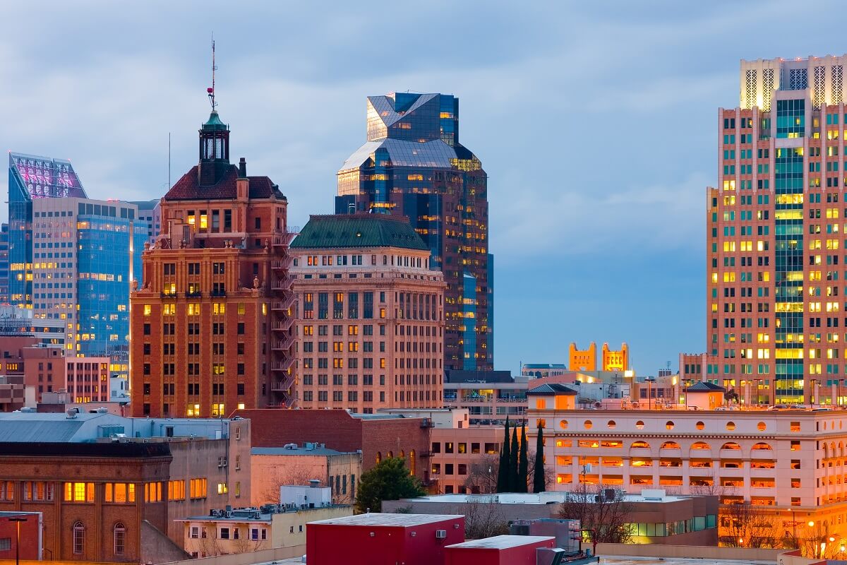 A cityscape at dusk featuring illuminated office buildings and skyscrapers under a partly cloudy sky.