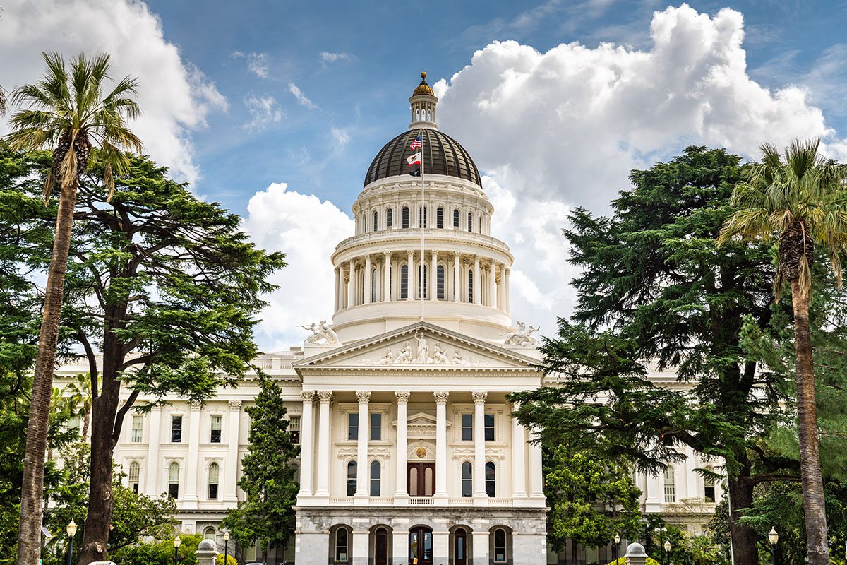 The California State Capitol building in Sacramento, featuring a white dome, columns, and surrounded by trees under a partly cloudy sky.