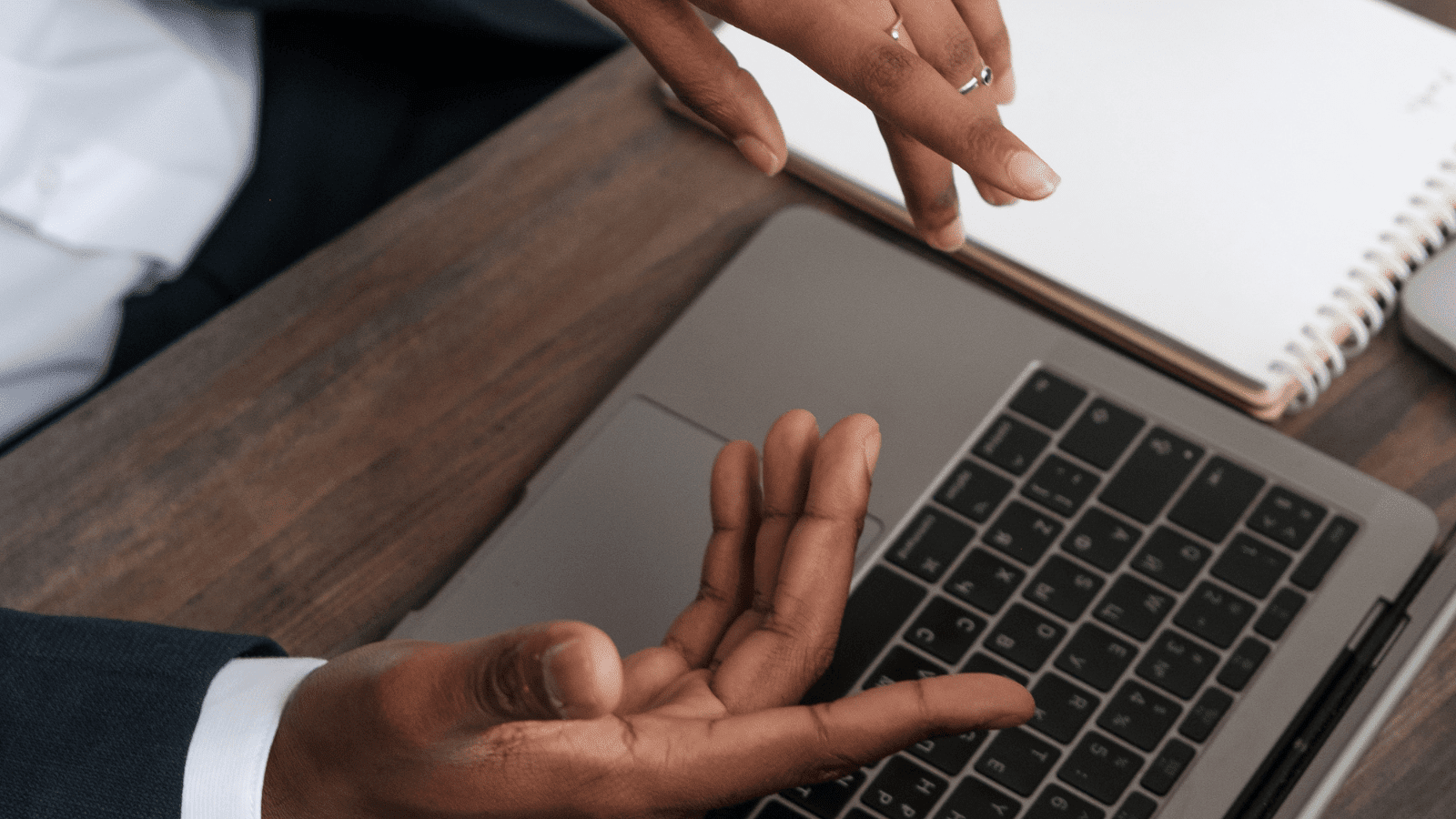 Two people gesture over a laptop keyboard on a wooden desk, with a notebook and pen visible nearby.