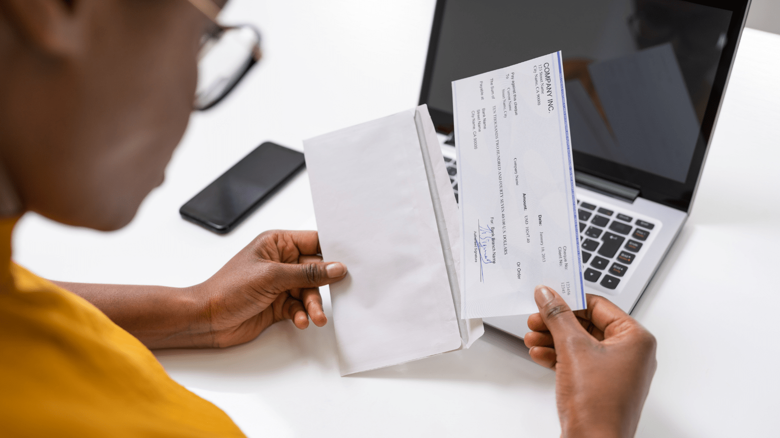 A person sits at a desk with a laptop, holding an envelope and a check, with a smartphone nearby.