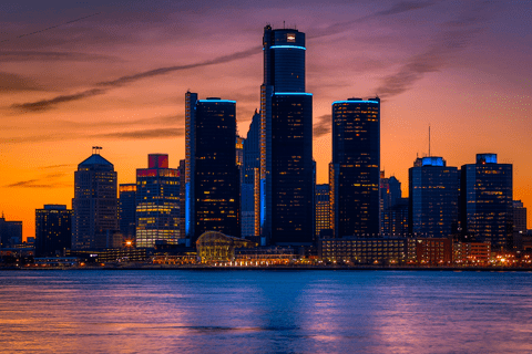 Downtown city skyline with tall buildings illuminated at dusk, seen across a river under a colorful sunset sky.