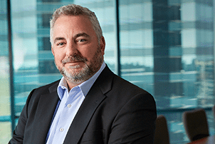 A man with gray hair and a beard wearing a suit jacket and light blue shirt sits in a modern office with large windows in the background.