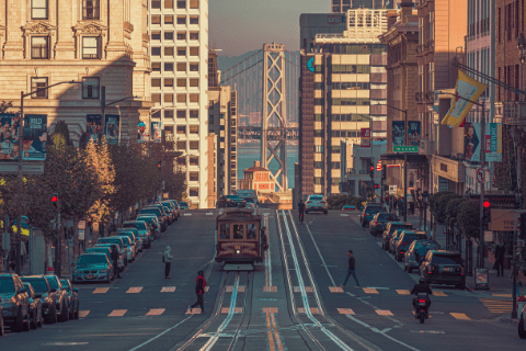 A steep city street in San Francisco with a cable car, parked cars, and the Bay Bridge visible in the background under clear skies.