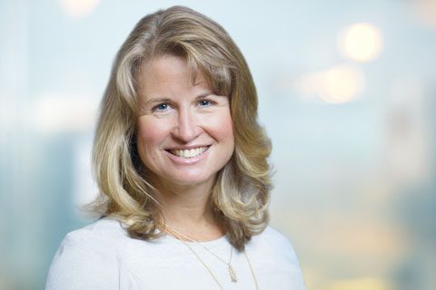 A woman with shoulder-length blonde hair, wearing a white top and a necklace, smiles at the camera against a softly blurred background.