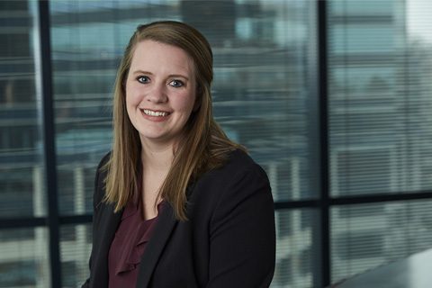 A woman with long brown hair wearing a blazer and blouse sits in front of large office windows with blinds and an urban background.