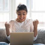 A person sits on a sofa, smiling and raising clenched fists in excitement while looking at a laptop.