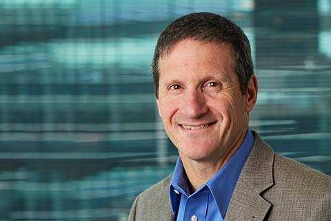 A man wearing a gray blazer and blue shirt smiles at the camera, standing in front of a blurred background with blue and green tones.