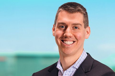 A man in a suit jacket and checked shirt smiles at the camera against a blurred outdoor background with a blue sky.