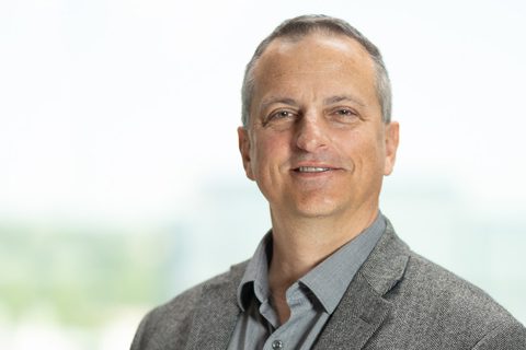 A middle-aged man wearing a gray blazer and collared shirt smiles slightly while standing in front of a blurred, light-colored background.