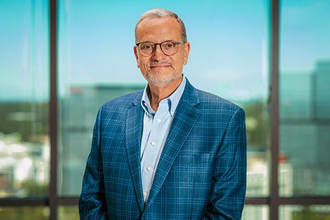 An older man with glasses and a gray beard wearing a blue plaid suit jacket and light blue shirt stands in front of large office windows with a cityscape view.