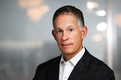 A middle-aged man with short gray hair wearing a dark suit and white shirt poses in front of a blurred office background.