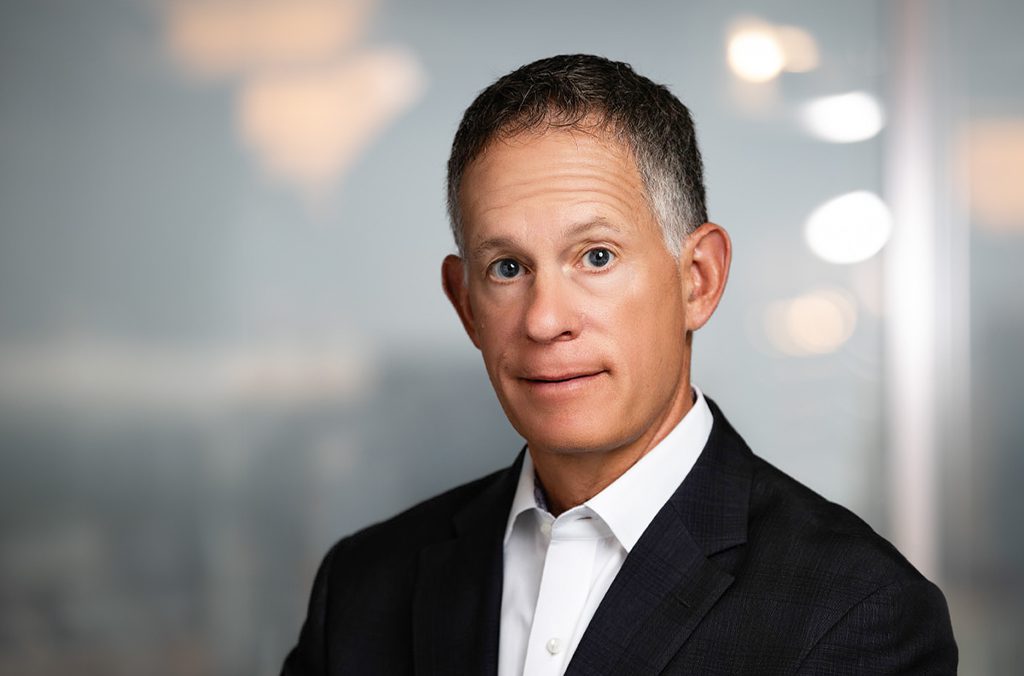 A man in a dark suit and white collared shirt poses for a professional portrait against a blurred, light-colored background.