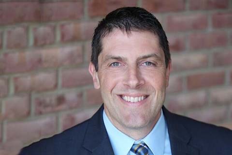 Man in a suit and tie smiling at the camera, standing in front of a brick wall background.