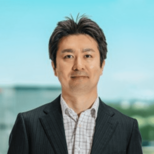 A man in a dark suit jacket and checked shirt stands outdoors with a blurred background of sky and buildings.