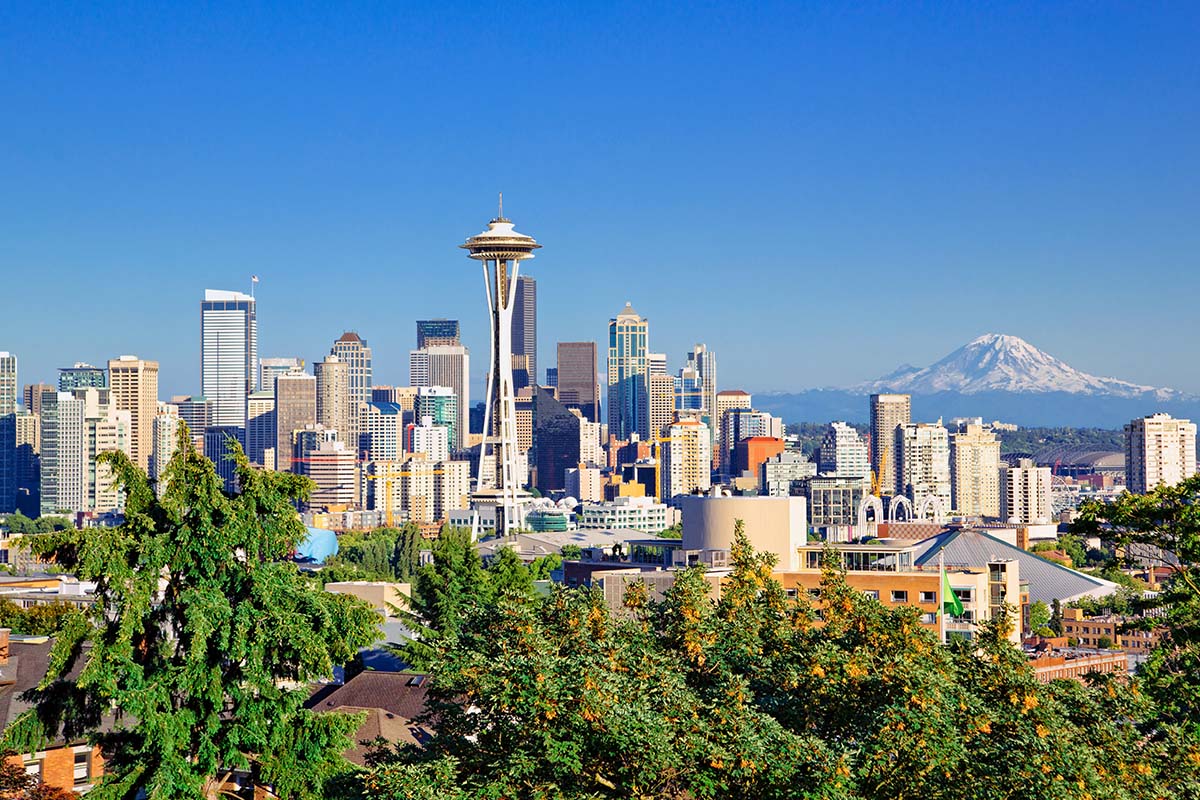 Seattle skyline with the Space Needle in the center, surrounded by skyscrapers, trees in the foreground, and Mount Rainier visible in the background under a clear blue sky.