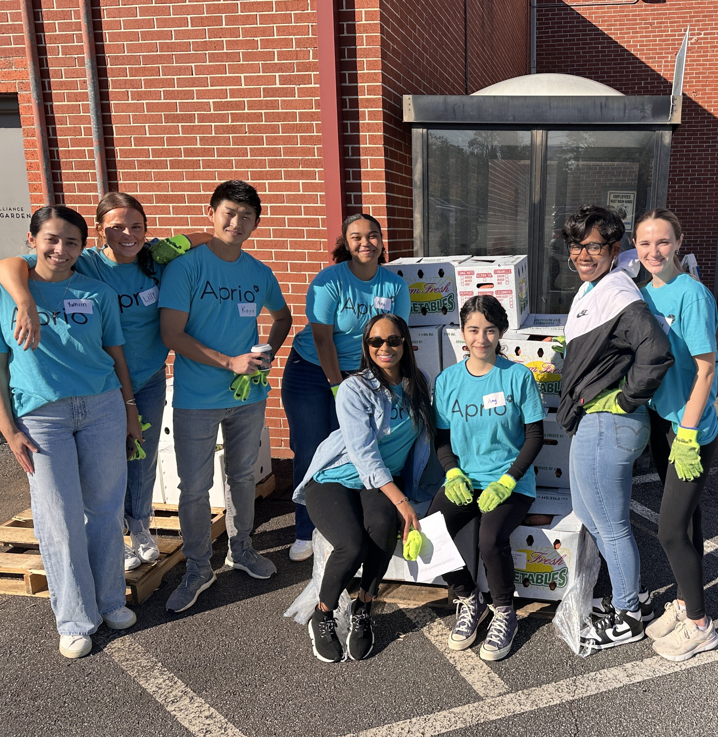 A group of eight people wearing blue Abria shirts and yellow gloves pose together outside in front of stacked boxes and a brick building.