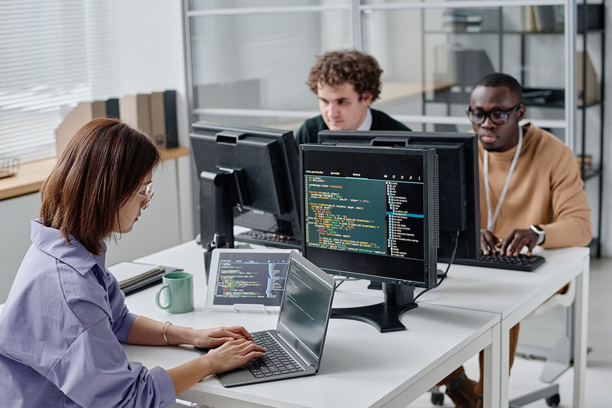 Three people work at a desk with computers displaying code, collaborating in a modern office with shelves and large windows.