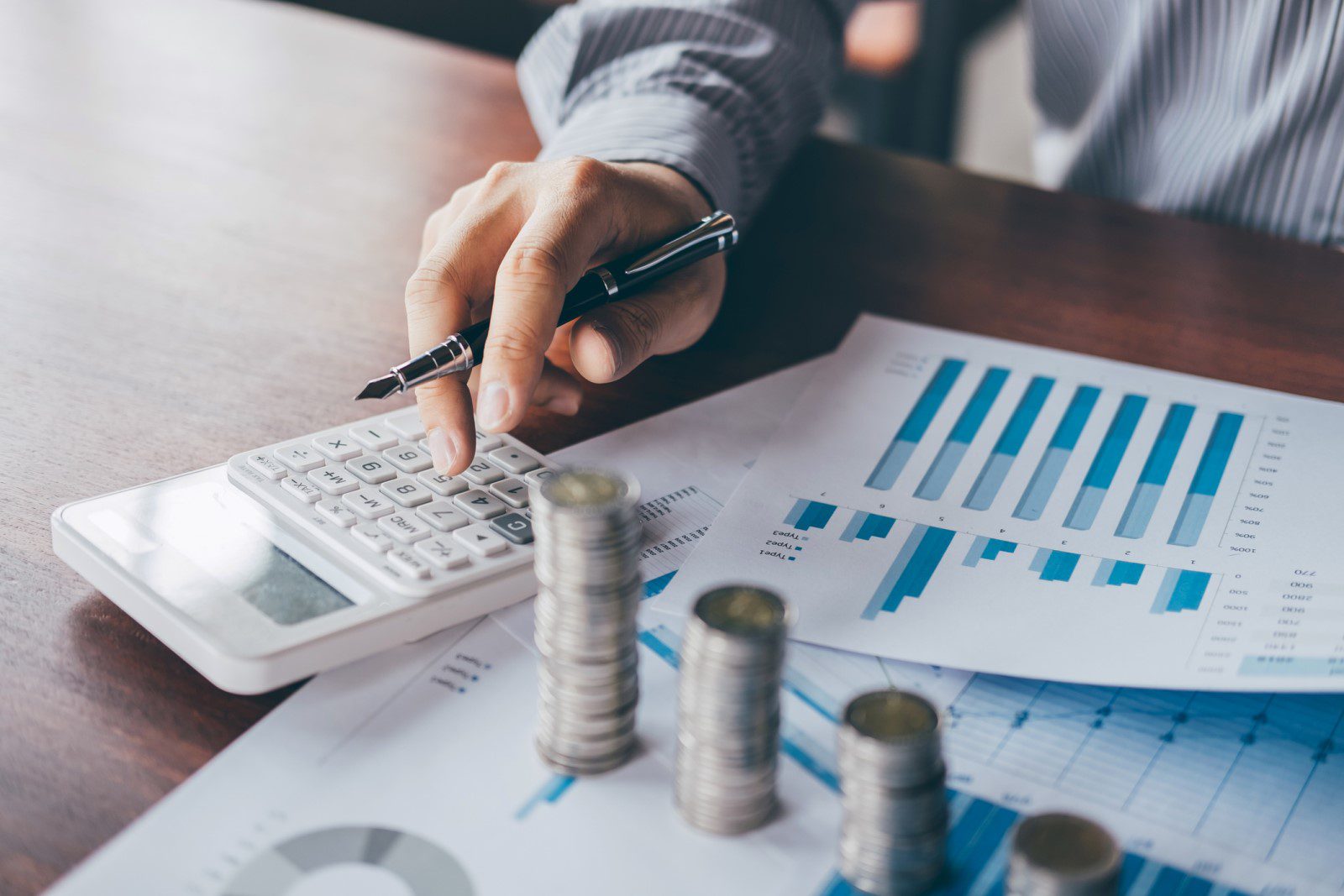 A person uses a calculator at a desk with stacked coins and financial charts, indicating calculations or financial analysis.