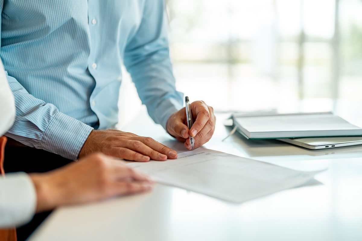 Two people stand at a desk as one person signs a document with a pen; an open laptop and papers are visible in the background.