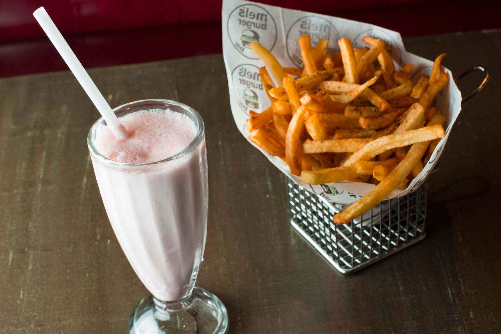 A glass of pink milkshake with a straw next to a metal basket filled with French fries on a wooden table.