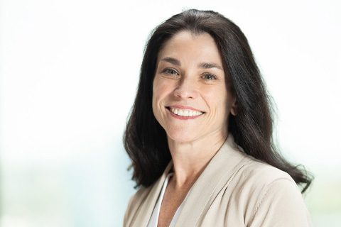 Woman with long dark hair wearing a beige blazer and white top, smiling at the camera against a bright, blurred background.