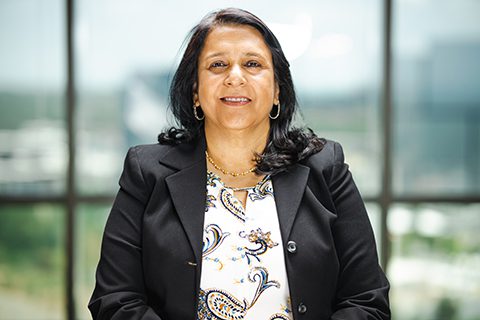 A woman with long dark hair wearing a black blazer and patterned blouse sits indoors in front of large windows.