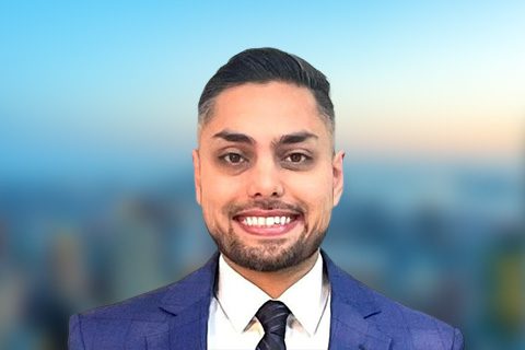 Dark haired beardedman in a blue blazer smiles in front of a bright window with a blurred city view