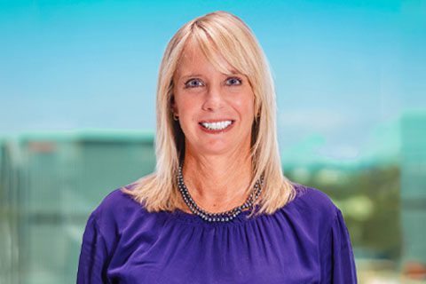 A woman with straight blonde hair wearing a purple blouse and beaded necklace smiles at the camera with a blurred cityscape background.