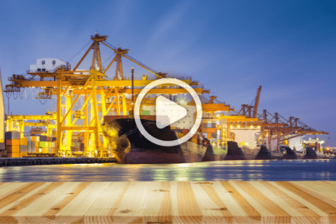 Shipping containers and cranes at a busy port are illuminated at dusk, seen behind a wooden surface in the foreground.