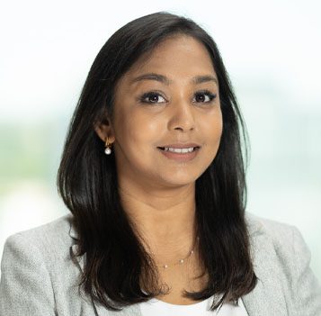 A woman with long dark hair wearing a light grey blazer, white top, and pearl earrings, poses in front of a blurred light background.