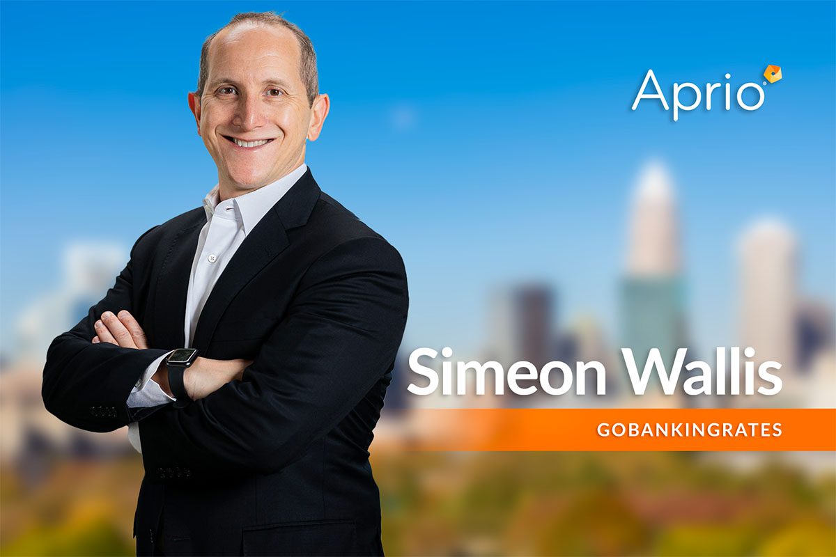 A man in a suit stands with arms crossed in front of a blurred cityscape background. The text reads Aprio, Simeon Wallis, and GOBANKINGRATES.