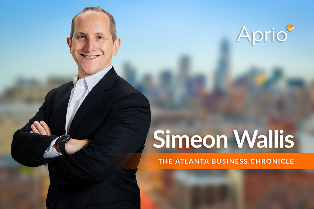 A man in a suit stands smiling with arms crossed in front of a blurred cityscape background. Text reads Aprio, Simeon Wallis, and The Atlanta Business Chronicle.