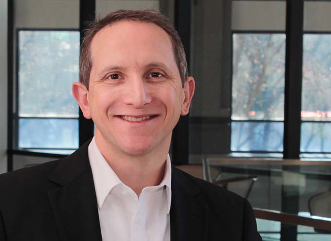 Man in a black suit jacket and white shirt smiling, standing indoors in front of large windows.
