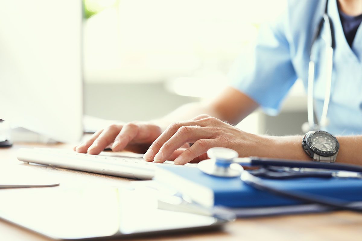 A person in medical scrubs types on a computer keyboard at a desk, with a stethoscope, clipboard, and documents nearby.